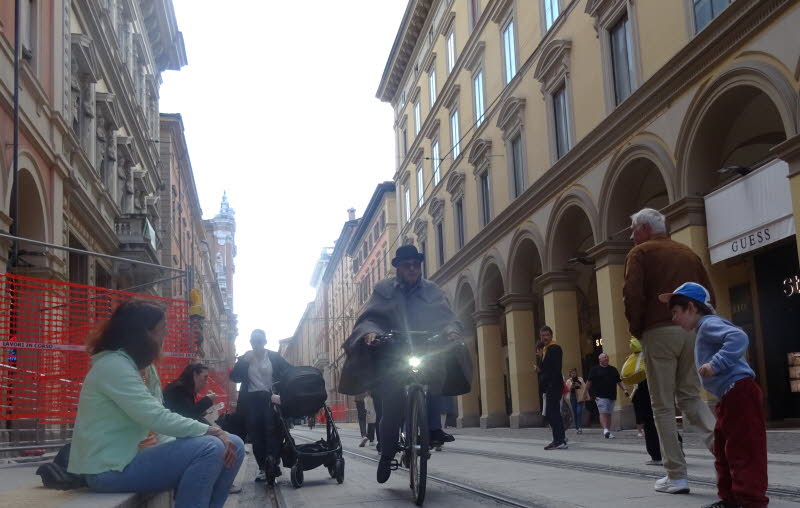Das Foto zeigt eine belebte Stra�enszene in Bologna mit historischer Architektur. Zentrale Figur ist eine Person auf einem Fahrrad mit hellem Frontlicht; mehrere Fu�g�nger, ein Kinderwagen und ein Kind sind im Bild. Am Ende der Stra�e ist ein Turm erkennb