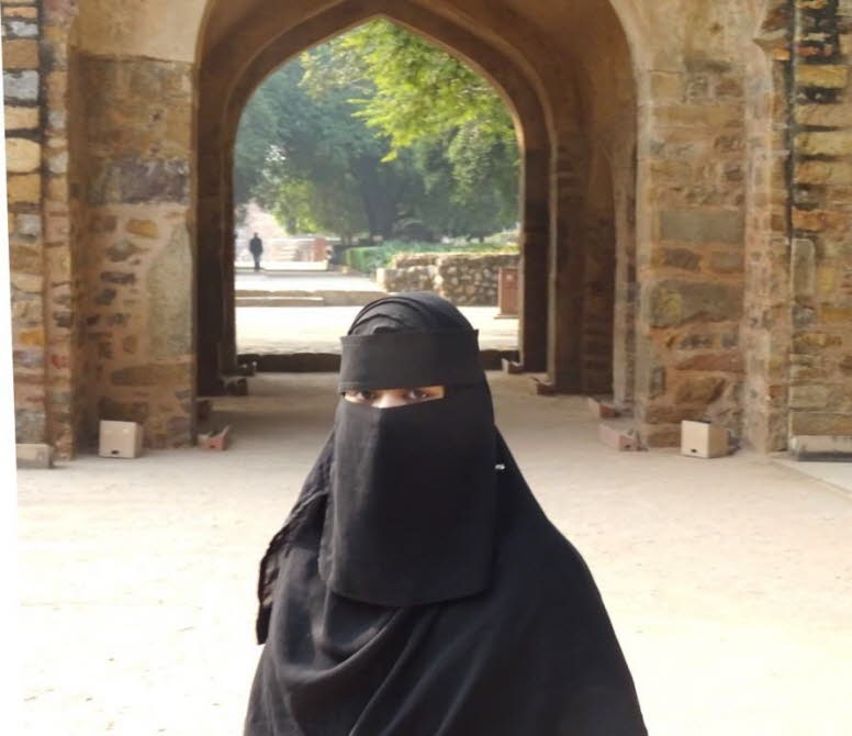 Das Bild zeigt eine Person, die eine schwarze Niqab und Abaya tr�gt, w�hrend sie durch einen historischen Steinbogen geht. Im Hintergrund sind weitere B�gen und eine baumges�umte Allee zu sehen. Die Kleidung der Person, bestehend aus Niqab und Abaya, ist 