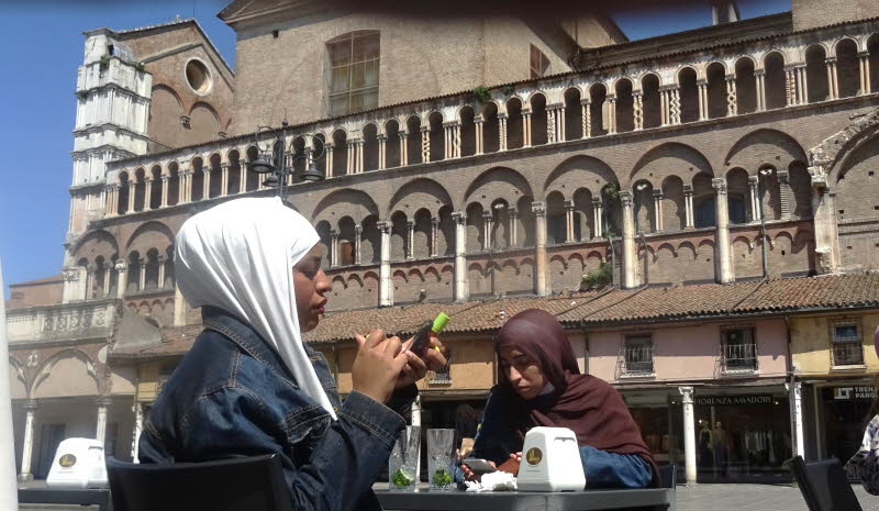 Fotografische Analyse der Szene: 1. Komposition & Bildaufbau: Zentrale Achse: Die beiden Personen sitzen nahezu mittig im Bild, wodurch eine stabile, ruhige Komposition entsteht. Architektur als Rahmen: Die romanische Fassade mit ihren Rundb�gen, S�ulen u
