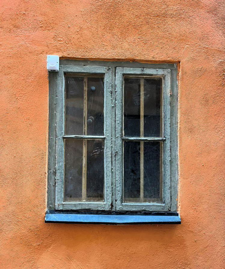 Das Bild zeigt ein klassisches Sprossenfenster mit grauem Holzrahmen, das tief in eine raue, orangefarbene Putzfassade eingelassen ist. Solche Gestaltungen sind typisch f�r traditionelle nordische Architektur, wie man sie beispielsweise in Stockholm finde