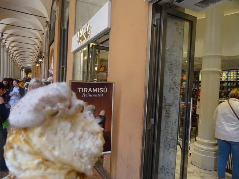 Auf dem Schild wird Tiramisu als Eiscreme-Variante beworben, beschrieben als "reinvented" (neu erfunden). Die Szene zeigt eine typische Eisdiele oder Gelateria.Solche italienischen Eiscaf�s bieten oft eine gro�e Auswahl an klassischen und kreativen Sorten