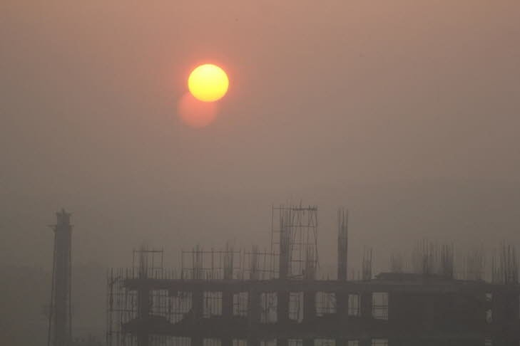 Das Bild zeigt einen Sonnenauf- oder -untergang �ber einer Baustelle in einem st�dtischen Gebiet in Delhi. Der Himmel ist aufgrund von Dunst oder Smog stark getr�bt, was zu der orange-rosa F�rbung f�hrt. Die Sonne erscheint als helle, klar definierte Sche