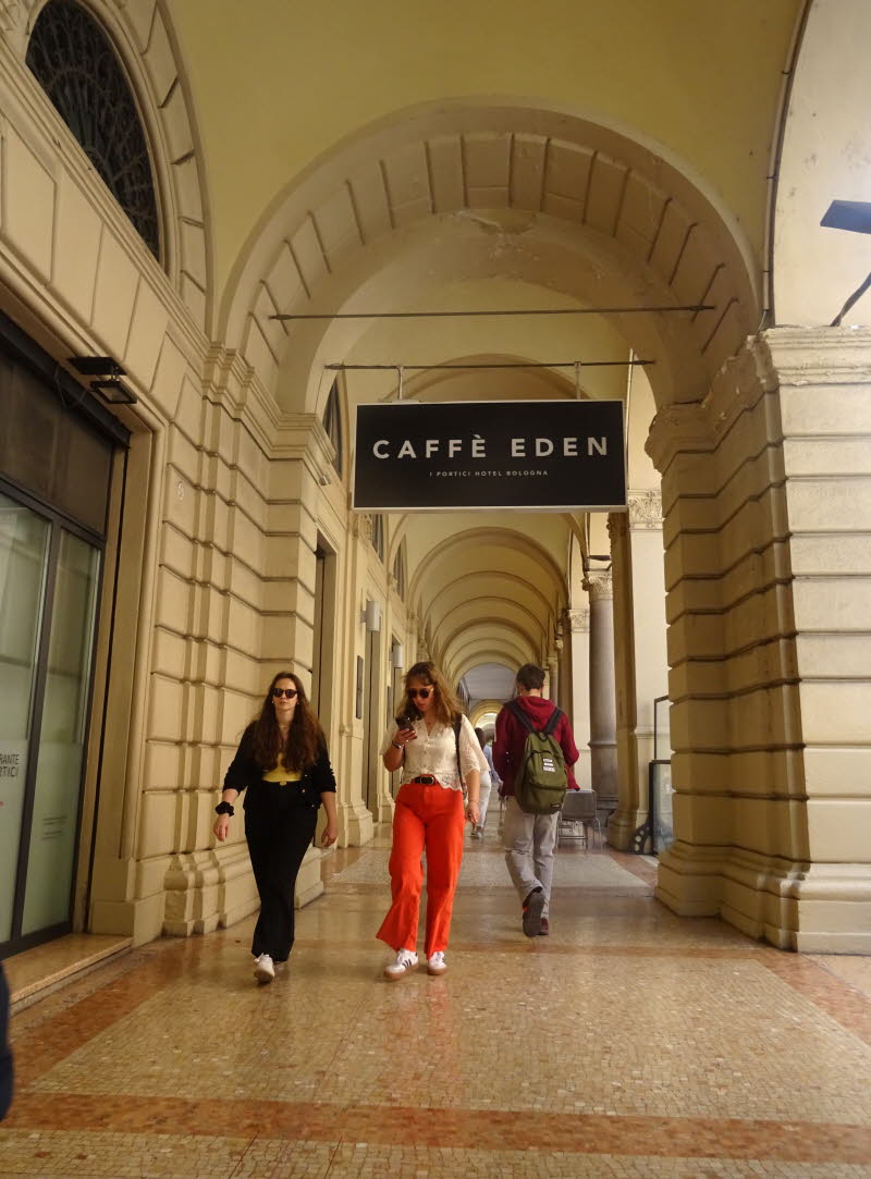 Ein �berdachter Arkadengang mit Tonnengew�lbe und rhythmisch gesetzten S�ulen.  Ein schwarzes Schild mit wei�er Schrift: CAFF� EDEN I PORTICI HOTEL BOLOGNA.  Drei Personen bewegen sich durch den Gang � zwei kommen dir entgegen, eine geht weg.  Der Boden b