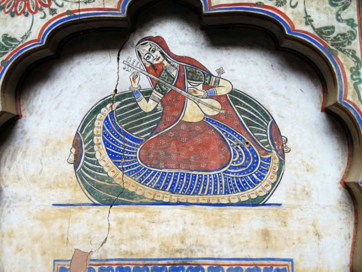 Das Bild zeigt ein traditionelles indisches Wandgem�lde, m�glicherweise in der Shekhawati-Region in Rajasthan. Es stellt eine Frau dar, die ein Saiteninstrument spielt, das einer Vina oder Sitar �hnelt. Die Malerei ist in einem Nischenbogen ausgef�hrt und