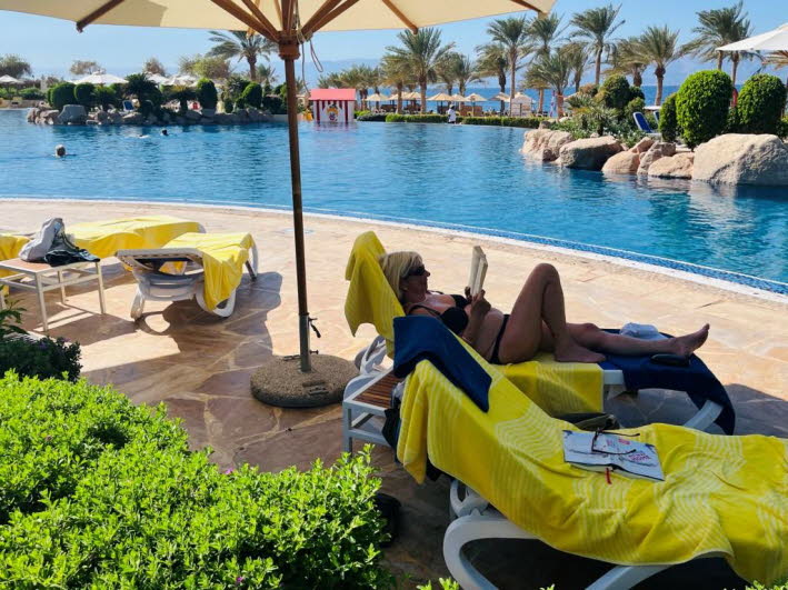 Die abgebildete Szene zeigt eine Person, die sich am Pool des M�venpick Resort & Spa Tala Bay Aqaba in Jordanien entspannt. Das 5-Sterne-Resort liegt direkt am Roten Meer in einem exklusiven Wohngebiet, etwa 15 Minuten vom Stadtzentrum Akabas entfernt. Es
