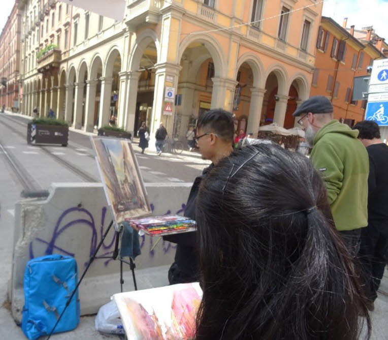 Das Bild zeigt eine Stra�enszene in Bologna mit Stra�enmalern bei der Arbeit, starker st�dtischer Architektur im Hintergrund und einer klaren Trennung zwischen Vordergrund (Maler, Staffelei, Material) und Hintergrund (Passanten, Geb�ude, Gleise). Die Szen