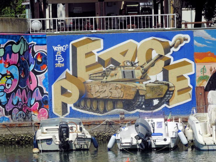 Dieses Wandbild zeigt einen beigen Kampfpanzer am Fluss Marecchia in Rimini, der von goldenen, dreidimensionalen Buchstaben umrahmt wird, welche das Wort "PEACE" (Frieden) bilden. Das Graffiti wurde im Jahr 2024 von einem K�nstler namens "MORIN" erstellt,