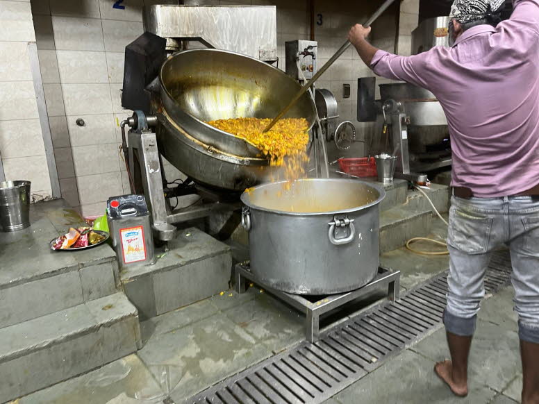 Das Bild zeigt die Essenszubereitung in einer Gro�k�che, wahrscheinlich in einem Gurdwara (einem Sikh-Gotteshaus), wo eine Mahlzeit namens Langar f�r die Gemeinschaft zubereitet wird. Ein Koch gie�t eine gro�e Menge einer gelben, gekochten Speise aus eine