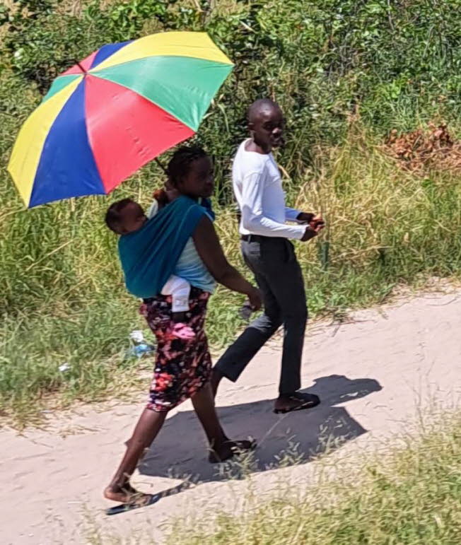 Das Bild zeigt eine Alltagsszene aus Namibia, in der eine Familie oder eine Gruppe von Personen zu Fu� auf einem unbefestigten Sandweg unterwegs ist.Kulturelle und allt�gliche Aspekte im Bild: Traditionelles Babytragen: Eine Frau tr�gt ein Kleinkind auf d