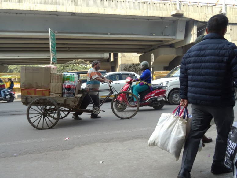 Das Bild zeigt einen Mann, der einen �berladenen Handwagen in einer belebten Stra�e in Alt-Delhi, Indien, zieht. Der Mann tr�gt eine blaue Jacke und schiebt den Wagen, der mit gro�en, in wei�es Material verpackten Ballen beladen ist. Die Szene spielt sich