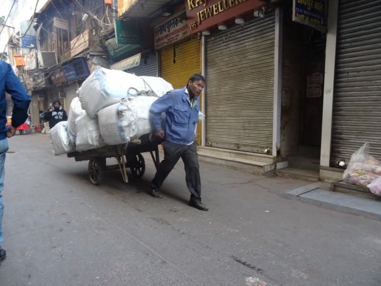 Das Bild zeigt eine belebte Stra�enszene, wahrscheinlich in Indien, mit mehreren Personen und Fahrzeugen: Ein Mann schiebt ein Lastenrad, das mit mehreren Kartons und Getr�nkeflaschen beladen ist. Eine Person mit Helm f�hrt einen roten Motorroller neben d