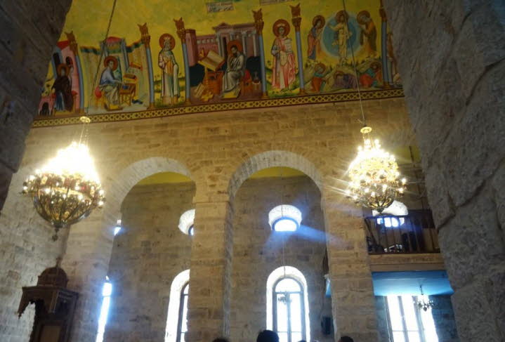 Die Kirche zeichnet sich durch rohe Steinmauern und mehrere B�gen aus. Mehrere Kronleuchter h�ngen von der Decke und beleuchten den Raum. Ein gro�es Wandgem�lde mit religi�sen Figuren und Szenen ist �ber den B�gen zu sehen. Menschen sind im unteren Teil d