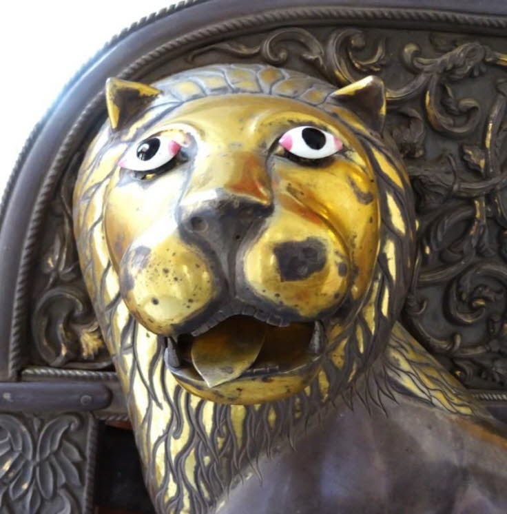Die Skulptur ist aus Metall, wahrscheinlich Messing oder einer Legierung, gefertigt und weist eine goldene Patina auf. Dieses L�wenmotiv ist Teil eines traditionellen Hathi Howdah (Elefantensitz), einer Art k�niglicher S�nfte, die im Museum ausgestellt wi