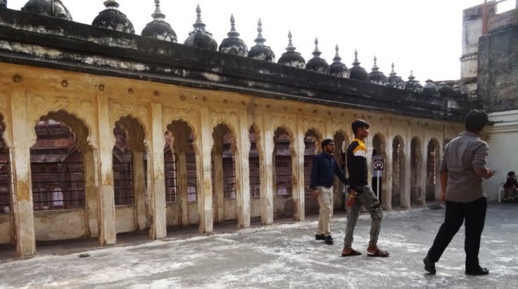 Auf dem Bild ist eine �berdachte Arkade in einem Innenhof des Mehrangarh Forts in Jodhpur, Rajasthan, Indien, zu sehen. Die Szene zeigt traditionelle Architektur und mehrere Personen, die sich im Bereich der Arkaden aufhalten. Architektonische Merkmale: B