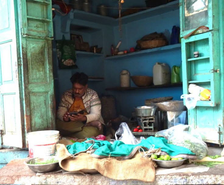 Das Bild zeigt einen Mann, der in einem kleinen, traditionellen Laden in Indien sitzt und sein Smartphone benutzt.