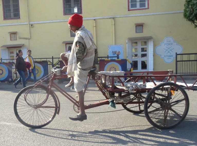 Das Bild zeigt einen Mann, der eine Fahrradrikscha (oder einen Lastenrikscha) in Jaipur, Rajasthan, Indien, f�hrt. Der Mann tr�gt traditionelle indische Kleidung, darunter einen roten Turban und eine Dhoti. Die traditionelle Kleidung der M�nner in Rajasth