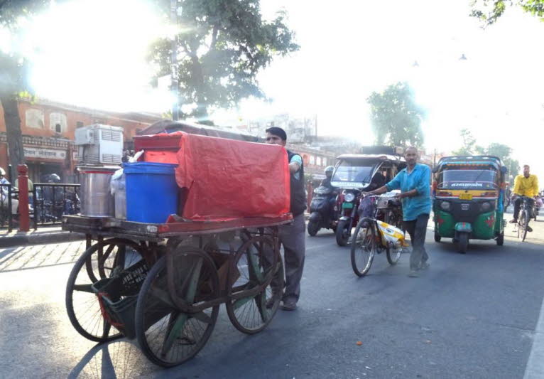Dies ist eine typische Stra�enszene aus einer Stadt in Indien, die das gesch�ftige Alltagsleben zeigt. Szene Details: Verkehr und Transportmittel: Die Stra�e ist belebt mit verschiedenen indischen Transportmitteln. Im Vordergrund befindet sich ein Verk�uf