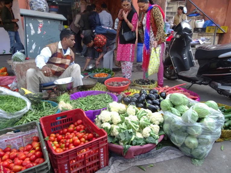 Das Bild zeigt einen gesch�ftigen Gem�semarkt im Freien in Jaipur mit mehreren Personen und einer Vielzahl frischer Produkte. Ein H�ndler sitzt hinter einer gro�en Auswahl an frischem Gem�se, darunter Tomaten, Blumenkohl, Auberginen, Bohnen und Kohl. Zwei