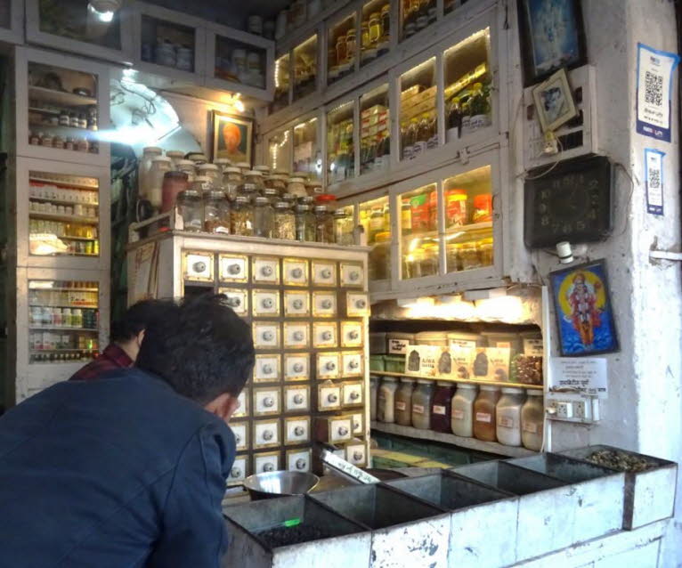 Das Bild zeigt das Innere eines traditionellen indischen Ladens, m�glicherweise eines Ayurveda- oder Gew�rzladens, mit einem Mann, der an der Theke einkauft. Der Laden verf�gt �ber zahlreiche Regale und Glasbeh�lter, die verschiedene Produkte wie Gew�rze,