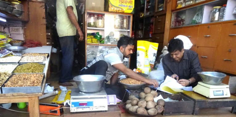 Das Bild zeigt einen gesch�ftigen Gew�rz- oder Nussmarkt, in Jaipur. Zwei M�nner sind aktiv mit dem Wiegen und Verpacken von Produkten besch�ftigt. Offene S�cke und Schalen mit N�ssen und Trockenfr�chten sind in den Regalen und auf den Tischen ausgestellt