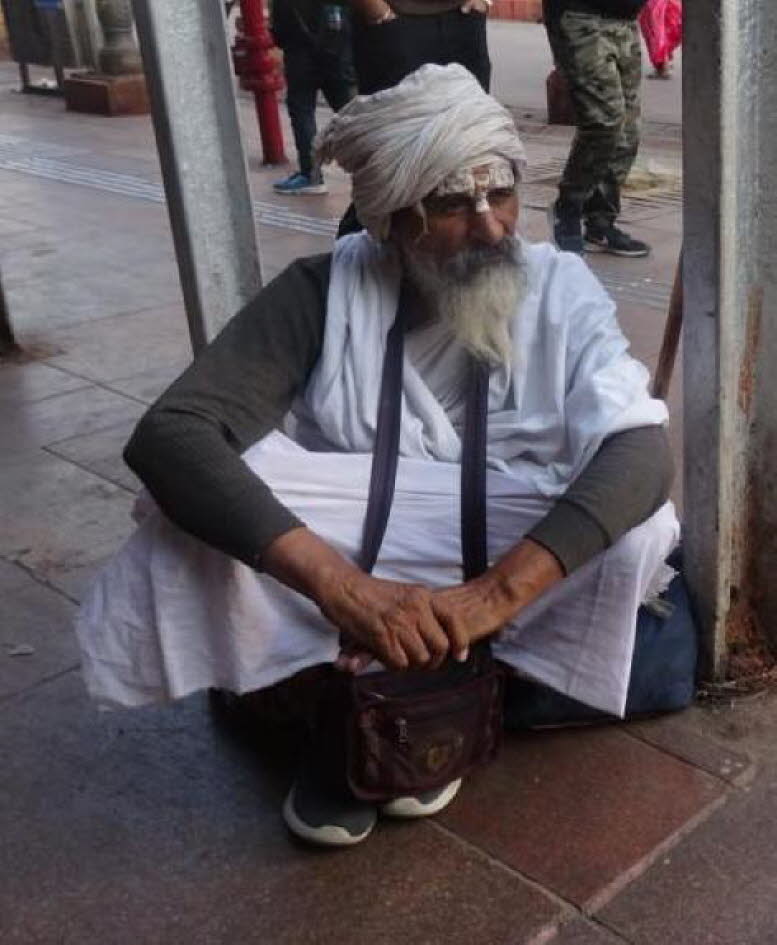 Das Bild zeigt einen sitzenden Mann, der traditionelle indische Kleidung tr�gt, vermutlich in einem Bahnhofsbereich in Indien. Der Mann tr�gt einen wei�en Turban und einen langen wei�en Bart. Er sitzt auf dem Boden, lehnt sich an einen Metallpfosten und h