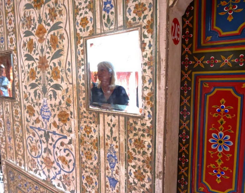 Die Abbildung zeigt eine Frau im Fort, die sich in einem Spiegel, der in eine reich verzierte Wand eingelassen ist, spiegelt. Die W�nde sind mit detaillierten, floralen Mustern in warmen Farben wie Orange, Gelb, Blau und Gr�n geschm�ckt. Die Verzierungen 