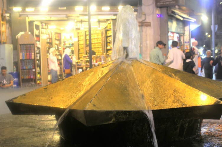 Belebter  Markt in Jordanien mit einem goldenen Springbrunnen im Vordergrund und einem Gesch�ft im Hintergrund. Der Springbrunnen hat eine einzigartige, eckige, goldfarbene Struktur. Im Hintergrund ist ein beleuchtetes Gesch�ft mit arabischem Text an der 