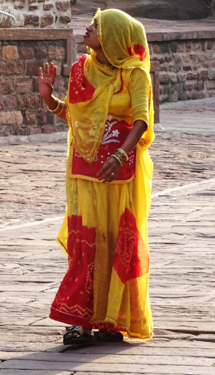 Die Frau tr�gt eine leuchtend gelbe Poshak (ein traditionelles Rajputi-Outfit) mit roten Akzenten, die typisch f�r die Region ist. Gelb symbolisiert oft Wissen oder Wohlstand, w�hrend Rot f�r Reinheit und eheliches Gl�ck steht. Sie tr�gt auch Armreifen al