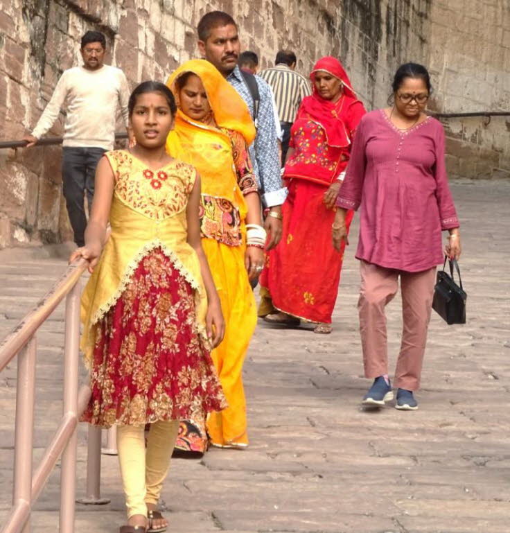 Die Abbildung zeigt eine Gruppe von Personen, darunter eine junge Frau oder ein M�dchen in einem gelb-roten Kleid und mehrere erwachsene Frauen in traditioneller indischer Kleidung, die eine Steintreppe hinaufgehen. 