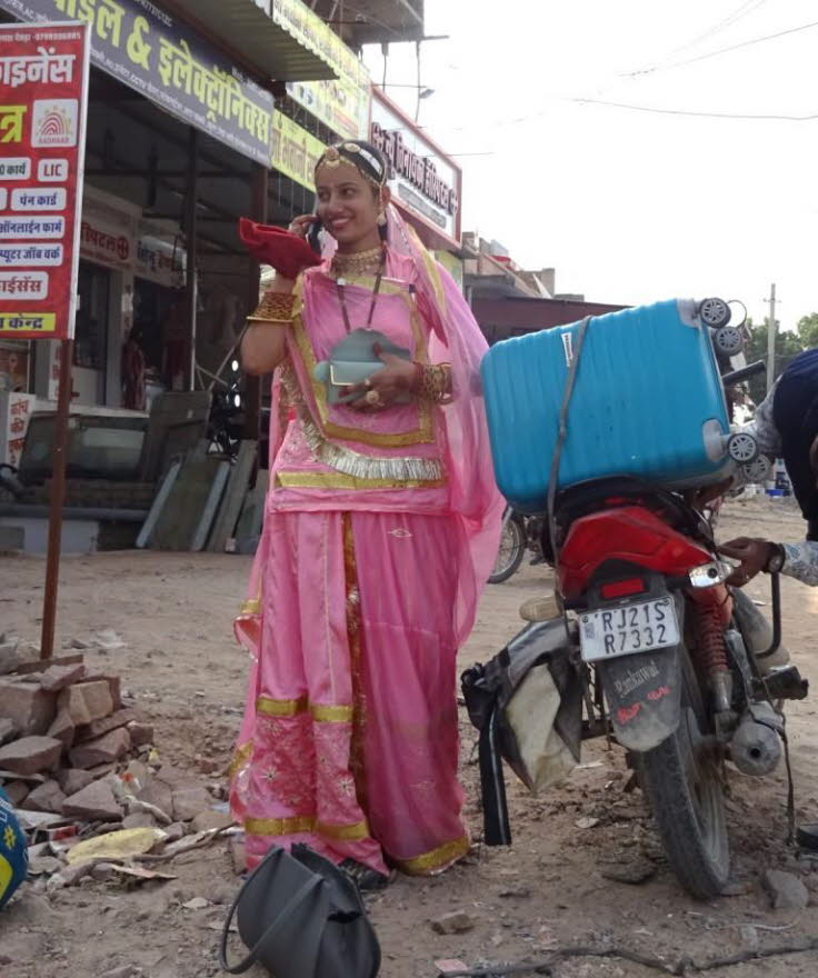 Die Frau tr�gt ein leuchtend pinkfarbenes traditionelles indisches Gewand, das mit goldenen Borten verziert ist. Dies k�nnte ein Ghagra Choli oder eine �hnliche regionale Tracht sein. Sie tr�gt auch traditionellen Schmuck und telefoniert. Die Szene spielt